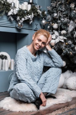 Şık örgü süveter giyen oldukça neşeli, genç, çekici sarışın kadın, Noel ağacının yanında, şık oyuncaklarla rahat bir oturma odasında dinleniyor. Mutlu kız model stüdyoda rahatlıyor..