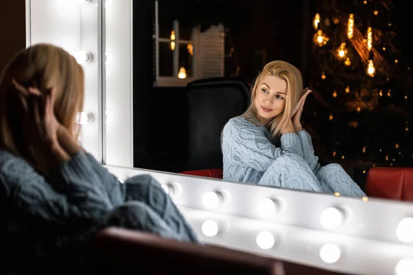 Mujer joven bastante glamour con un pelo rubio se sienta en una habitación con un árbol de Año Nuevo con luces brillantes y se ve en un espejo vintage con lámparas blancas. Hermosa chica modelo de moda a la espera de Navidad . — Foto de Stock