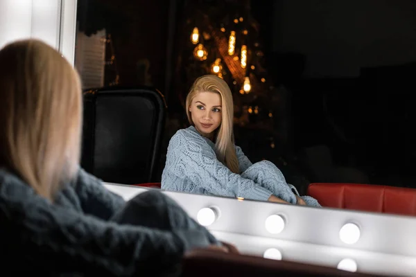 Trendy young woman in stylish knitted clothes sits and looking in a vintage white mirror in a room with Christmas decorations. Fashionable cute girl model is posing in the studio. Happy New Year. — 스톡 사진