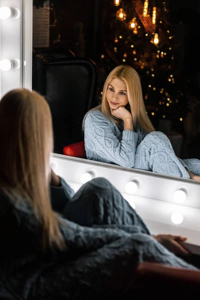 在古老的镜子旁边，坐着一位穿着蓝色针织服装的年轻貌美的女模特。 可爱的女模特儿晚上在一个有圣诞树的房间里休息。 背景色. — 图库照片