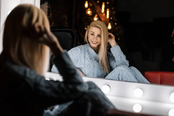 Attraktiv ung kvinna i snygga stickade kläder sitter och ler gulligt nära en vintage vit spegel i ett rum med nyårsdekorationer. Glada flicka modell vilar i studion. Gott nytt år. — Stockfoto