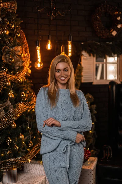 Elég divatos fiatal nő, elegáns kötött öltönyben áll és édesen mosolyog a nappaliban egy karácsonyfával, füzérekkel, fényekkel és ajándékokkal. Vonzó boldog lány vár karácsonyra. — Stock Fotó
