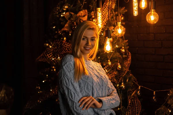 Feliz mujer joven atractiva en un elegante traje azul de punto posando en una habitación en el fondo de un árbol de Año Nuevo con luces y guirnaldas brillantes y hermosas. Chica feliz con una linda sonrisa en el interior . —  Fotos de Stock