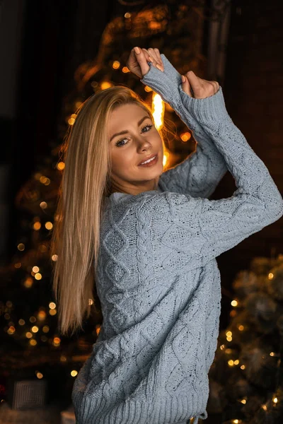 Joyful jovem mulher em um terno azul de malha na moda com cabelo loiro posando uma sala de estar com uma árvore de Natal à noite. Menina bonito feliz está de pé e sorrindo no estúdio com decoração de Ano Novo — Fotografia de Stock