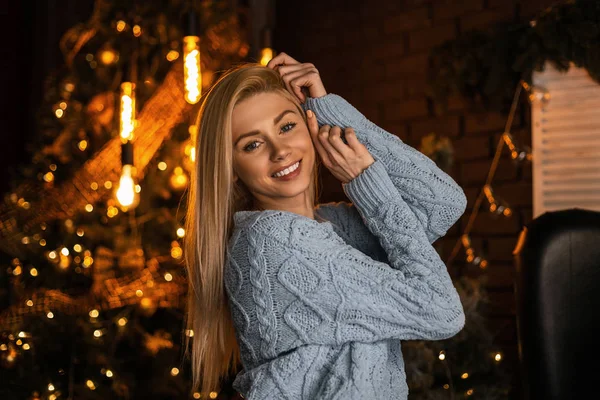 Retrato de uma jovem mulher feliz atraente com um belo sorriso em um terno de malha na moda no fundo de uma árvore de Natal festiva com guirlandas vintage. Menina elegante bonito posando dentro de casa . — Fotografia de Stock