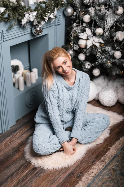 Pretty young woman in a fashionable knitted blue suit with blond hair with cute smile posing in a room with a Christmas tree. Attractive girl is sitting and smiling in the studio with New Year\'s decor