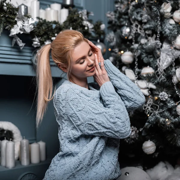 Joven mujer rubia glamorosa alegre en un suéter de punto de moda es relajarse en una acogedora sala de estar cerca de un árbol de Navidad y una chimenea vintage. Chica feliz modelo de moda se relaja en el estudio . —  Fotos de Stock