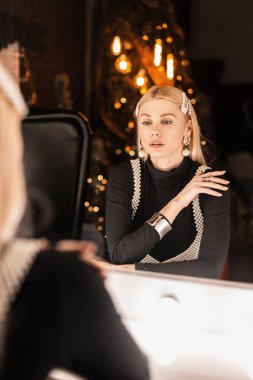 Siyah tişörtlü, siyah tokalı, saç tokalı çekici genç bir kadın karanlık oturma odasında lambaları olan antika bir aynanın yanında oturuyor. Şirin kız stüdyoda Noel ağacıyla rahatlıyor..