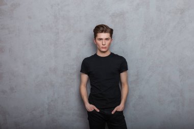 Modayı takip eden bir saç stili olan şık siyah kotlu, şık bir tişört giyen Avrupalı genç bir adam, odadaki gri bir duvarın yanında duruyor. Kapalı mekanda genç giysili yakışıklı hippi.