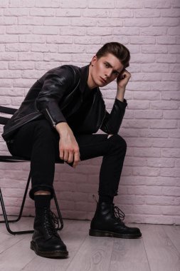Son moda deri ayakkabılı, modaya uygun siyah elbiseli genç bir adamın oldukça şık bir modeli stüdyodaki eski bir tuğla duvarın yanındaki bir sandalyede dinleniyor. Çekici hippi çocuk. Gençlik modası.