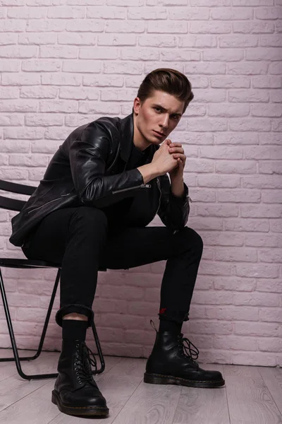 Fashion model of a serious young man in a vintage leather black jacket in stylish clothes in fashionable leather youth boots sitting on a chair near a pink brick wall indoors. Cool american guy.