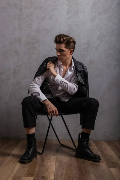 スタジオのヴィンテージの壁の近くの椅子に座ってスタイリッシュな黒いブーツでファッショナブルな服の髪型とトレンディな若いハンサムな男のヒップスター クールなセクシーな男がリラックスします 男性のためのファッション服 — ストック写真