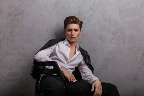 Young handsome man hipster in elegant white shirt in a fashionable leather black jacket is sitting on a chair near a gray vintage wall indoors. Attractive trendy guy model is resting in the studio.