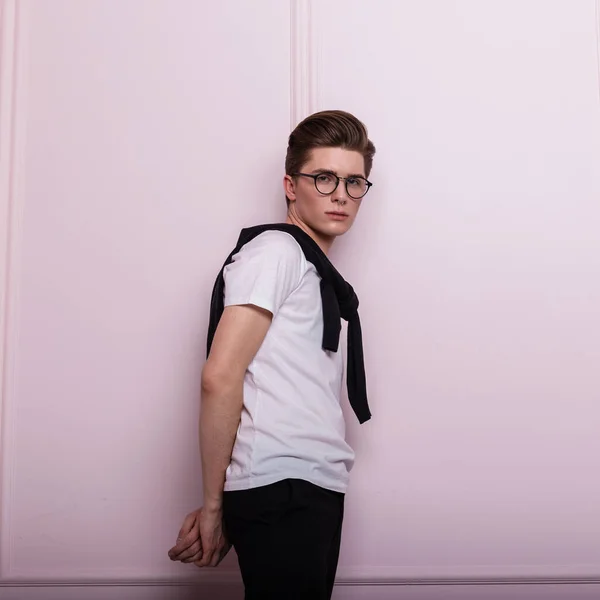 English handsome young man hipster in white stylish clothes with fashionable hairstyle in a glasses posing in room near a pink wall. Modern attractive guy fashion model in casual wear in the studio.