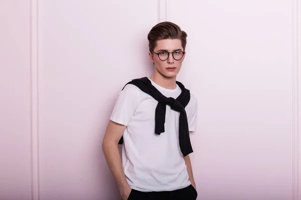 Pretty cute young man hipster in glasses in fashionable clothes is resting near a vintage pink wall in the studio. Elegant guy model with stylish hairstyle with pierced nose in trendy wear indoors.