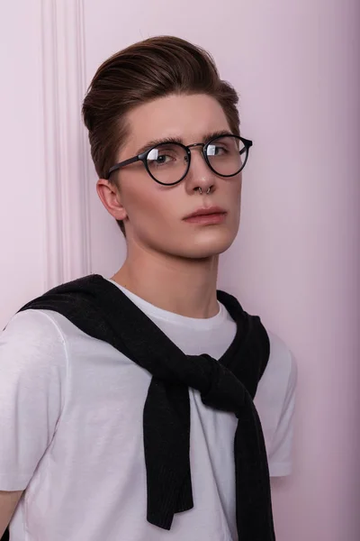 ピンクのヴィンテージの壁を背景にカーディガンで白いTシャツに鼻ピアスをした流行のメガネできれいな健康な肌を持つハンサムな若いヒップスター男のスタイリッシュなスタジオの肖像画 アメリカ人の少年 — ストック写真
