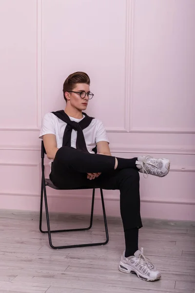 Young stylish man model in glasses in fashionable youth black-white clothes in trendy leather sneakers posing sitting on a vintage chair near a pink wall in the studio. Attractive hipster guy. Fashion