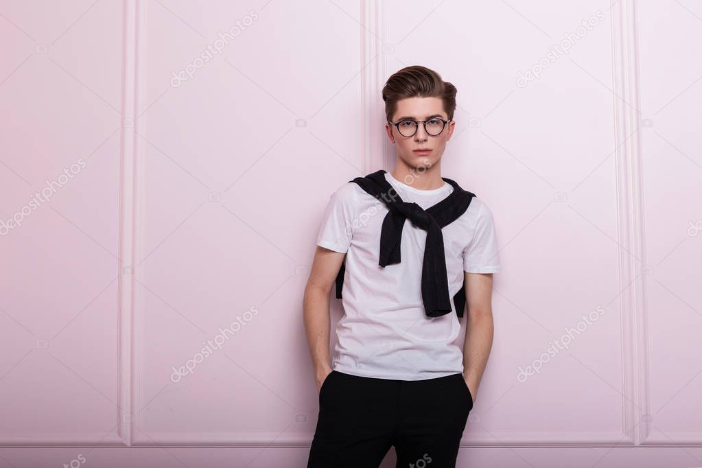 Fashionable attractive young man model in vintage glasses with a hairstyle in a trendy white t-shirt in black jeans with a nose piercing posing near a pink wall in the studio. Elegant hipster guy.