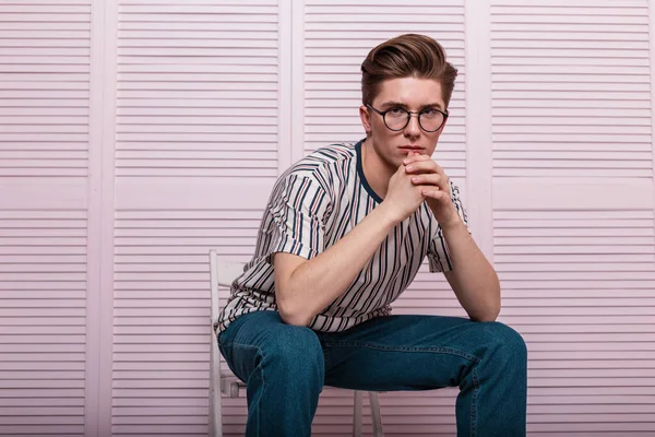 Young fashionable man with a stylish hairstyle in a fashion striped T-shirt in vintage glasses posing on a chair near a wooden pink wall in the room. Modern hipster guy in trendy youth wear indoors.