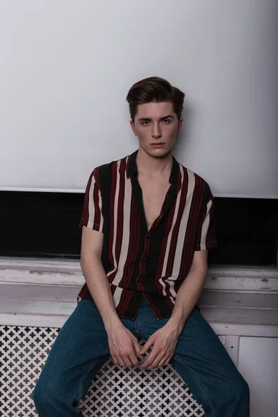 European serious young man in an elegant striped shirt in blue stylish jeans is resting near a white vintage window in the room. Attractive guy is a fashion model indoors. Fashionable clothes for men.