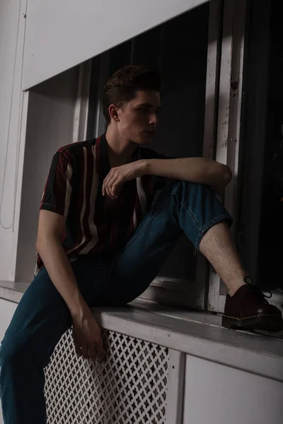 流行の髪型のヴィンテージジーンズのスタイリッシュな縞模様のシャツを着たファッショナブルな若い男が部屋の窓の近くの窓の上に休んでいます 魅力的な男のファッションモデルは屋内でリラックス — ストック写真