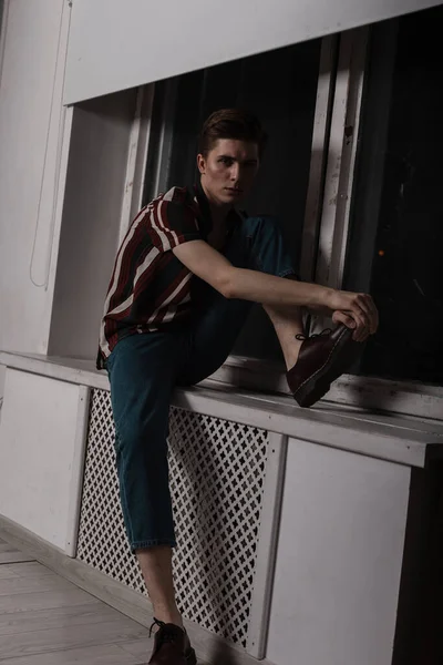 流行の髪型のヴィンテージジーンズを着たスタイリッシュなストライプのシャツを着たかなりかわいい若い男が窓の近くの窓の上に屋内に休んでいます ファッションモデルのハンサムな男リラックスでザ部屋 — ストック写真
