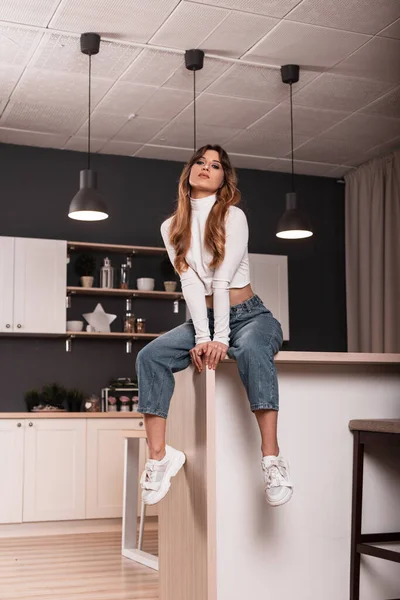 Fashionable sexy young woman model in a fashionable white top in blue vintage jeans in trendy sneakers sits on the kitchen table in the cook room. Stylish beautiful girl is resting in the studio.