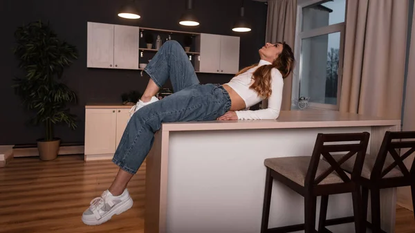 Pretty stylish young woman with red hair in fashionable clothes in stylish gym shoes sexually lies on a vintage kitchen table indoors. Trendy girl fashion model relaxes in a cook room.