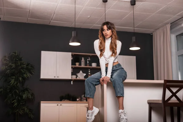 Attractive slim young woman model in a fashion white top in vintage jeans in trendy sneakers sexually sits on the kitchen table in the cook room. Beautiful fashionable girl is resting indoors.