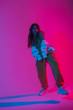Moda spor ayakkabılarıyla şık giyinen genç kadın manken modern bir stüdyoda çok renkli renklerle dans ediyor. Çekici kız dansçı stüdyoda neon ışıkla dans etmekten hoşlanıyor..
