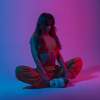 Moda pantolonlu, modaya uygun giyinmiş, stil diskinde çok renkli bir renk olan bir odada yoga yapan atletik genç bir kadın. Moda kıyafeti giyen profesyonel bir kız, parlak mavi ışıklı bir odada oturuyor.