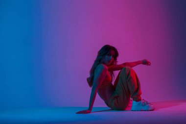 Moda mankeni, şık gençlik kıyafetleri içinde güzel, güzel, güzel bir kadın. Muhteşem, mavi bir ışığı olan bir odada dinleniyor. Modern moda kız stüdyoda neon renkli simlerle oturuyor..