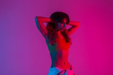 Şık bir bluzu olan, belinde kapüşonlu ve muhteşem kırmızı ışıkla içeride poz veren seksi, genç bir kadın. Modern güzel kız mankeni çok renkli bir odada düz saçlar yapıyor.