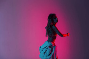 Belinde kapüşonlu şık bir kapüşonlu seksi genç bayan dansçı açık pembe ışıkla içeride dans ediyor. İnce vücutlu, uzun saçlı bir kız disko stilinde dans etmekten hoşlanıyor..