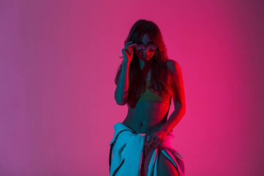 Moda kıyafetleri içinde oldukça modern genç hippi bir kadın göz kamaştırıcı pembe ışığı olan bir odada şık bir güneş gözlüğü takıyor. Seksi vücutlu güzel bir kız. Disko tarzı neon renkli bir evde poz veriyor..