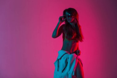 Şık gençlik kıyafetleri içinde modaya uygun genç kadın modelleri, pembe-kırmızı arka planlı kapalı mekanlarda moda güneş gözlüklerini düzeltiyor. Modern hippi kız disko tarzında renkli ışıklarla stüdyoda poz veriyor..