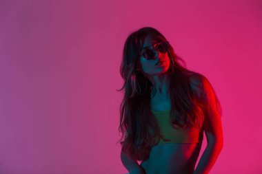 Havalı, uzun saçlı, modaya uygun giyinmiş genç bir hippi pembe bir arka planda bir odada poz veriyor. Modayı takip eden güneş gözlüklü, parlak pembe ışıklı bir kız stüdyoda duruyor.