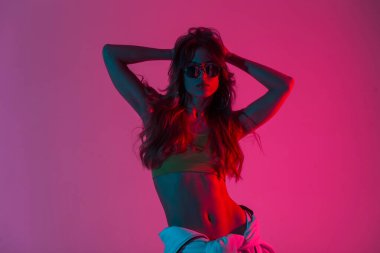 Güneş gözlüklü, seksi bir bluzu olan, uzun saçlı ve parlak kırmızı ışıktan hoşlanan genç model stil sahibi bir kadın. Çok renkli diskosu olan bir odada çekici bir hippi kız..