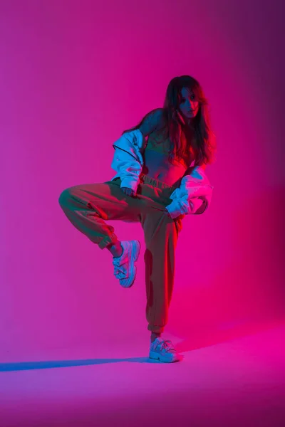 明るいピンクの紫外線の部屋に1本足でポーズを取るスニーカーで流行のパンツでスタイリッシュなトップで若い女性ダンサー ネオンマルチカラーで室内で踊るクールな現代的なスタイリッシュな女の子 — ストック写真