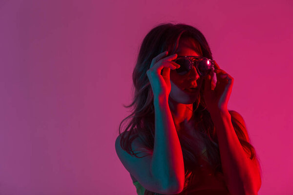 Портрет молодая женщина в верхней части с сексуальными губами с длинными волосами ставит на модные солнцезащитные очки в комнате с неоновым розовым цветом background.Modern хипстер девушка в помещении с ярко-красным удивительным светом. Крупный план
