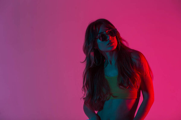 Крутая молодая женщина-хипстер с длинными волосами в модном топе позирует в комнате на розовом фоне. Сексуальная девушка в триумфальных солнцезащитных очках в трико стоит в студии с ярким неоновым розовым светом
