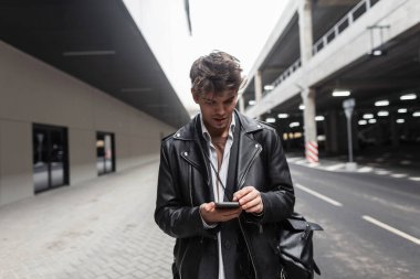 Deri ceketli, saç stili sırt çantalı, modern modacı genç adam sokaklarda telefonu elinde tutuyor. Moda kıyafetleri içinde çekici bir hippi ayakta durup akıllı telefona bakar..