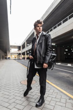 Moda modası geçmiş siyah günlük giysili modern genç hippi yol kenarında poz veriyor. Şık, modern kentsel adam modeli, şık bir deri ceketli, bol pantolonlu, açık havada botlu. Sokak modası.