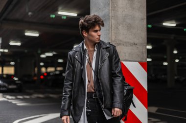 İş adamı hippi, saç stili modaya uygun, büyük beden deri ceketli sırt çantalı, park yerinde bir sütunun yanında dinleniyor. Profesyonel erkek modeli. Amerikan modası kıyafetleri.