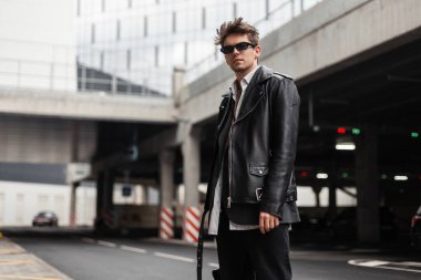 Klasik güneş gözlüklü, deri ceketli, kotlu modern model bir adam açık otoparkta duruyor. Şehrin sokaklarında gezen seksi Amerikalı havalı hippi. Retro biçimi.