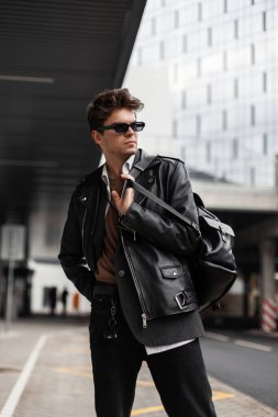 Siyah güneş gözlüklü, modaya uygun siyah ceketli çekici bir genç adam şehirde şık bir sırt çantasıyla poz veriyor. Yakışıklı Amerikalı hippi çocuk. Yeni koleksiyon erkek giyim