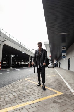 Modayı takip eden güneş gözlüklü, modaya uygun deri ceketli, klasik çizmeli Amerikalı genç hippi şehirde modern bir binanın yanındaki fayansta yürüyor. Açık havada çekici bir adam..