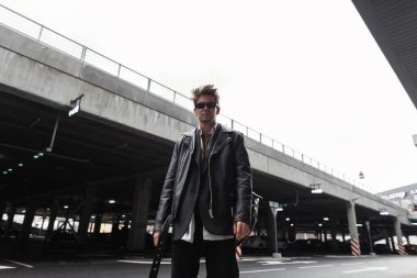 Havalı, modaya uygun, güneş gözlüklü, modaya uygun, modaya uygun siyah deri ceketli, modaya uygun bir genç hippi şehirdeki klasik bir binanın yanında duruyor. Sokaktaki ciddi modern adam..