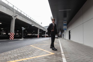 Siyah deri ceketli, botlu, güneş gözlüklü modern stil sahibi genç hippi otoparkın yanındaki sokakta yürüyor. Şehirde modaya uygun giyinen yakışıklı bir adam modeli..