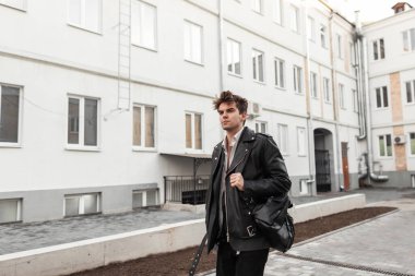 Moda derisi ceketli yakışıklı genç adam şık siyah bir sırt çantası ve modaya uygun bir saç stili ile klasik evlerin yanında sokakta yürüyor. Çekici Amerikalı hippi bahar günü yürüyüşe çıkmış..